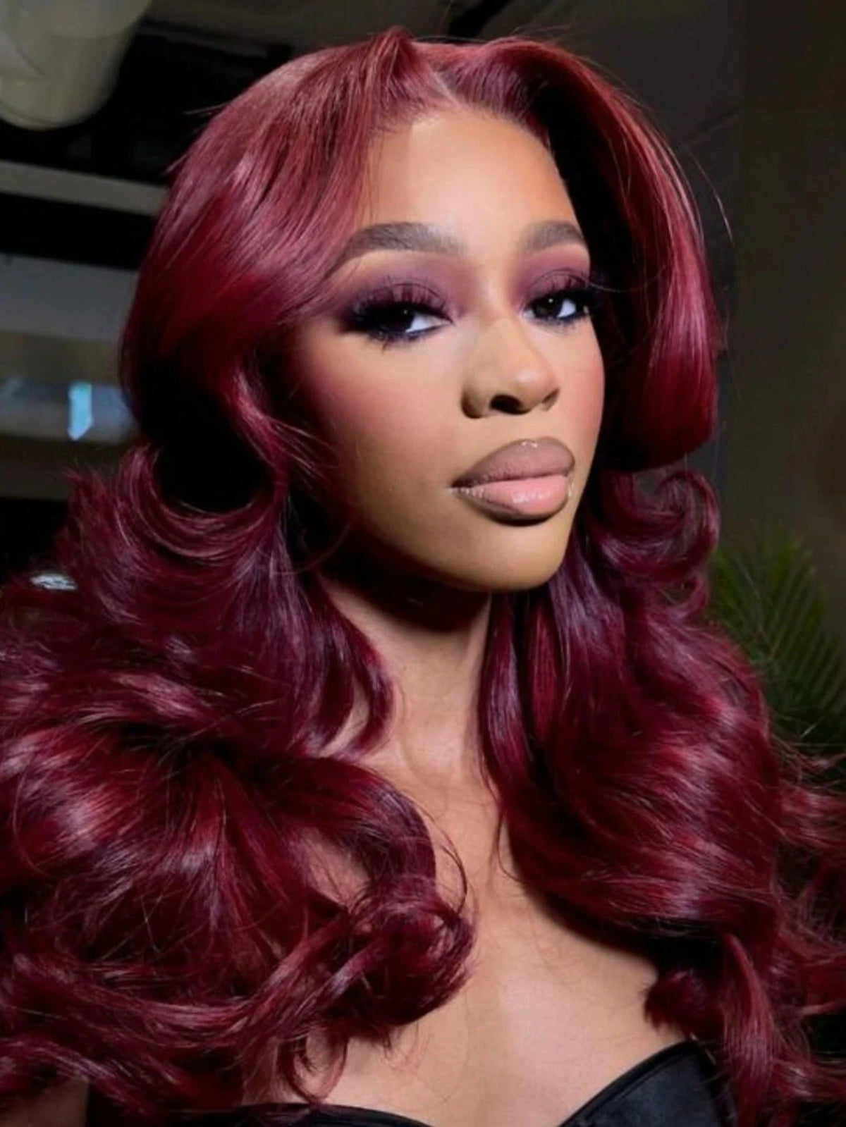 A close-up portrait of a woman with long, wavy burgundy hair wearing makeup.