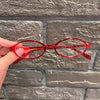 Une main tient une paire de lunettes de mode ovales rouges contre un mur de briques grises.