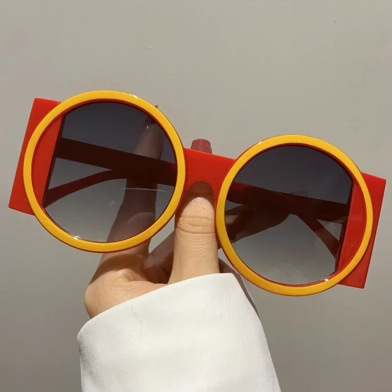 A hand holds a pair of round double color fashion sunglasses with orange and red frames.