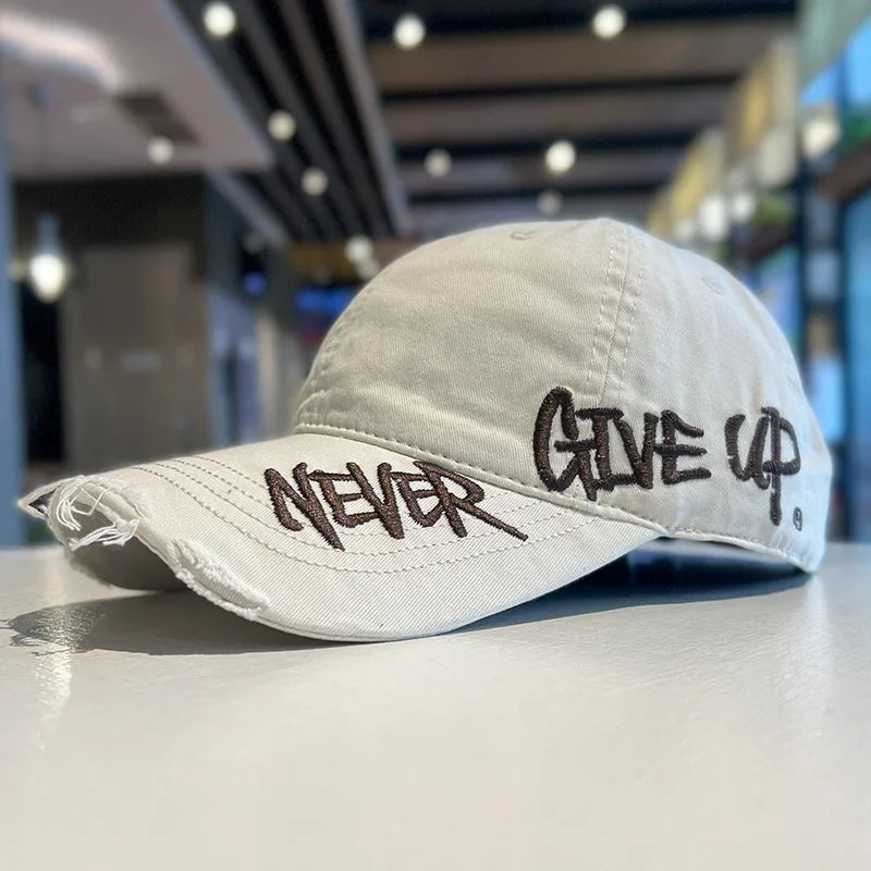 A light beige baseball cap with embroidered text that reads NEVER GIVE UP resting on a smooth surface.