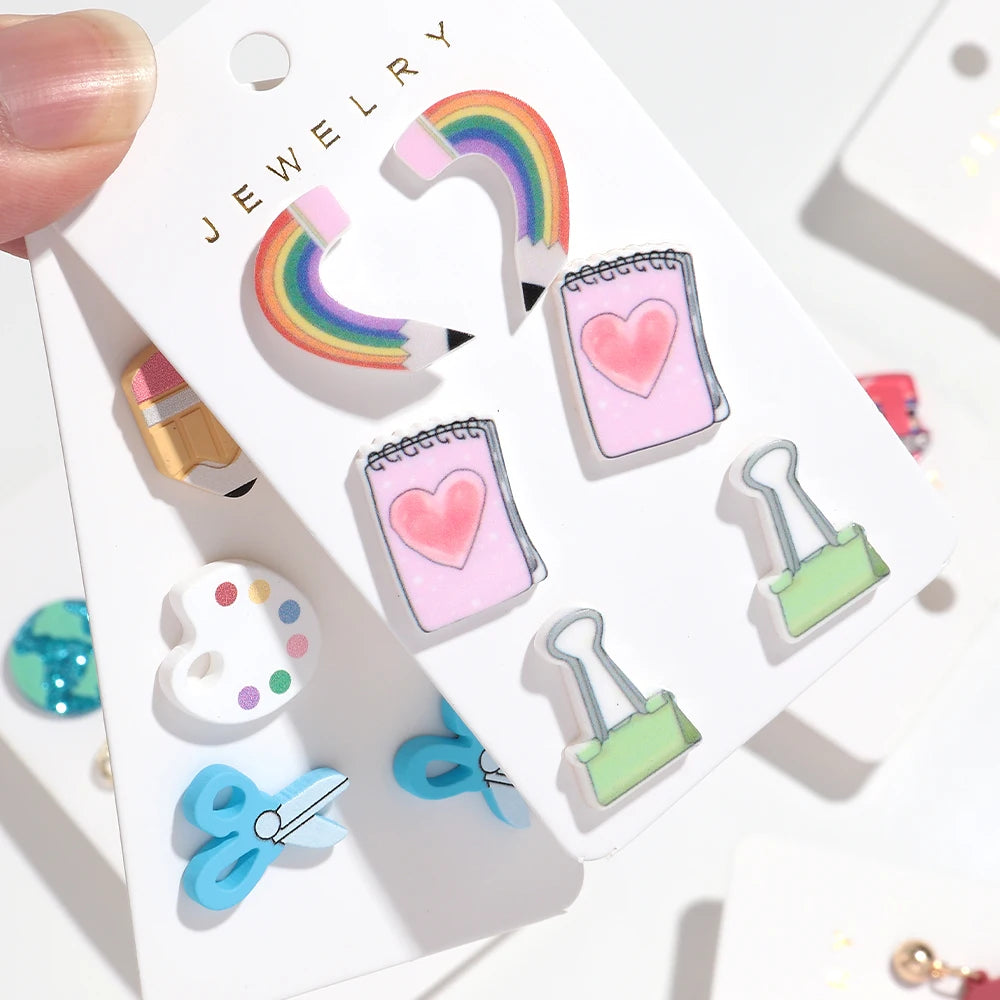 Colección de pendientes de gota en forma de lápiz y corazón de colores, exhibidos en una tarjeta blanca.