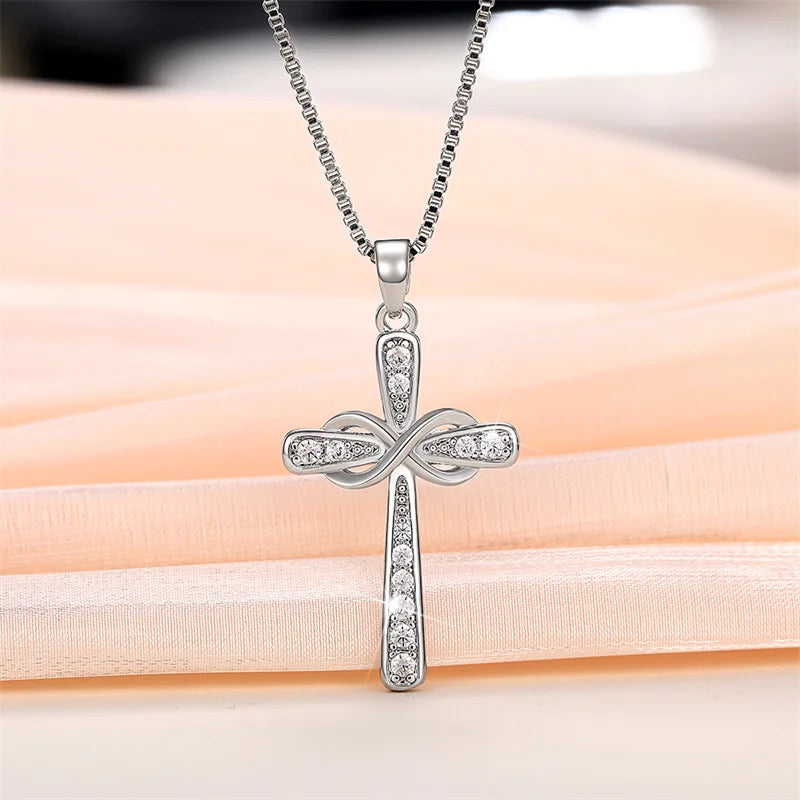 A silver cross pendant necklace with a detailed design is displayed on a pink fabric background.