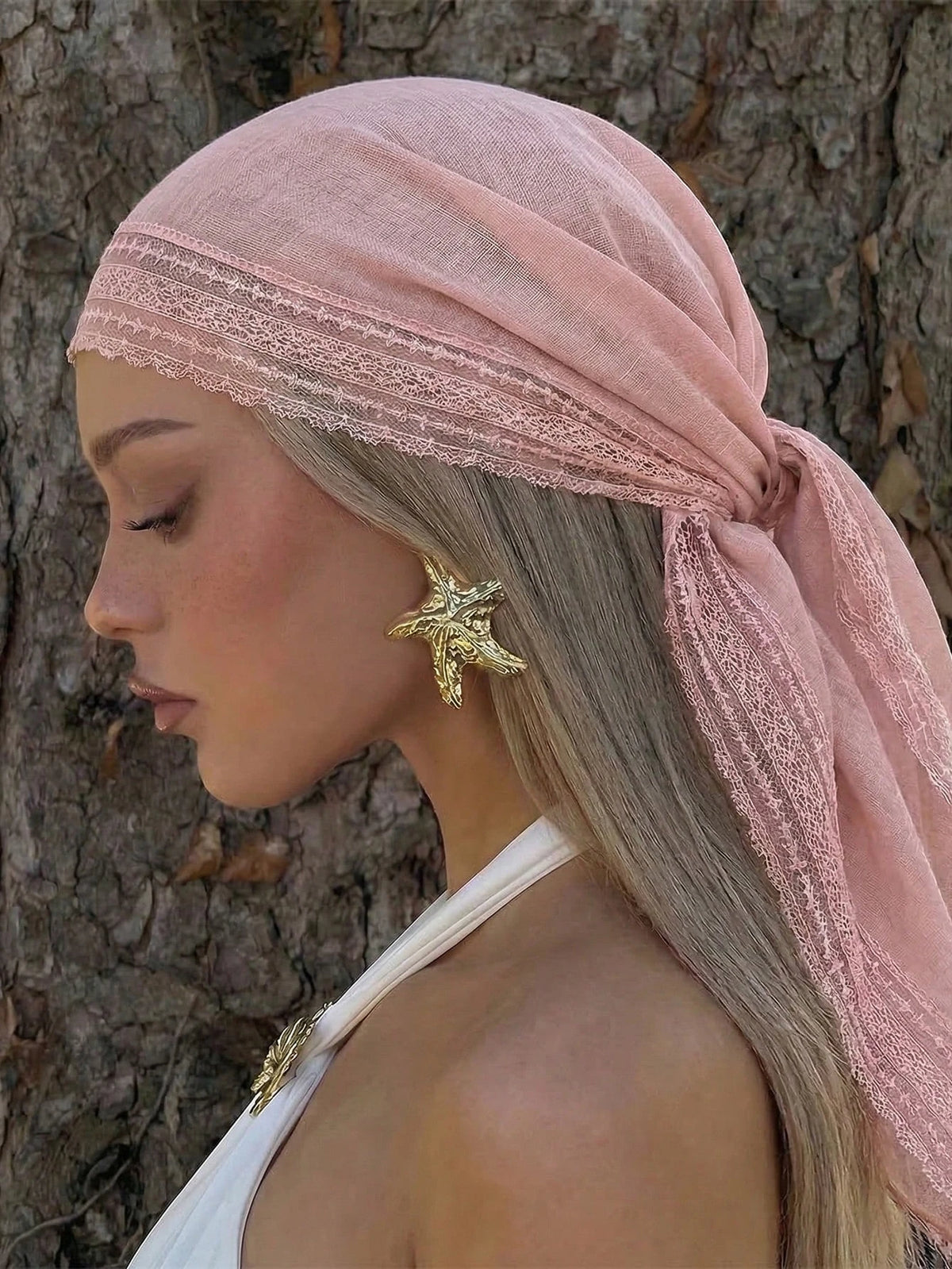 A woman wearing a dusty rose sheer lace trim shawl draped over her head and hair against a tree trunk.