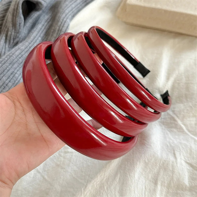 A hand holds a stack of glossy red PU leather fashion headbands against a light fabric background.