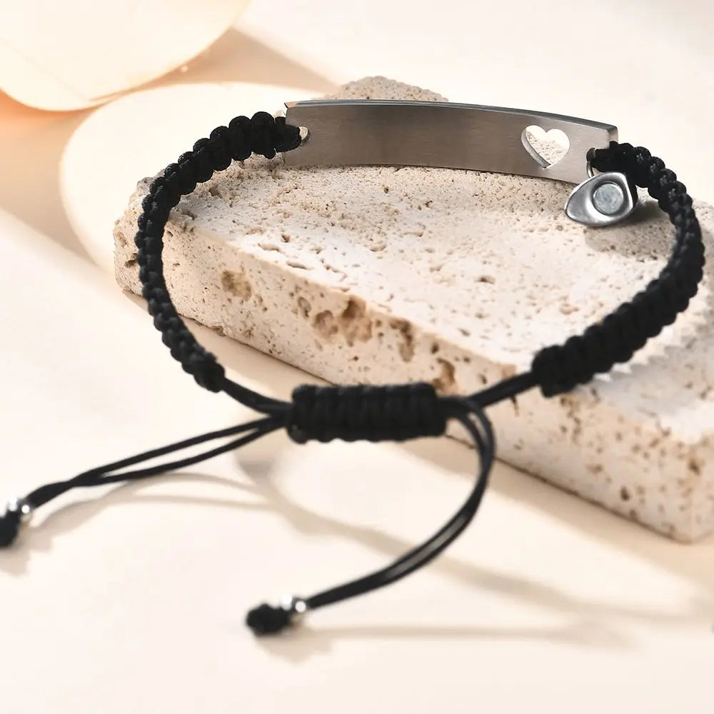 Maßgeschneidertes Edelstahl-Paararmband-Set mit Silberstab und schwarzem Schnur mit Herzanhänger auf hellem Stein