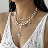 A close-up of a woman wearing a layered necklace featuring pearls, starfish, and shell charms against a white crochet top.