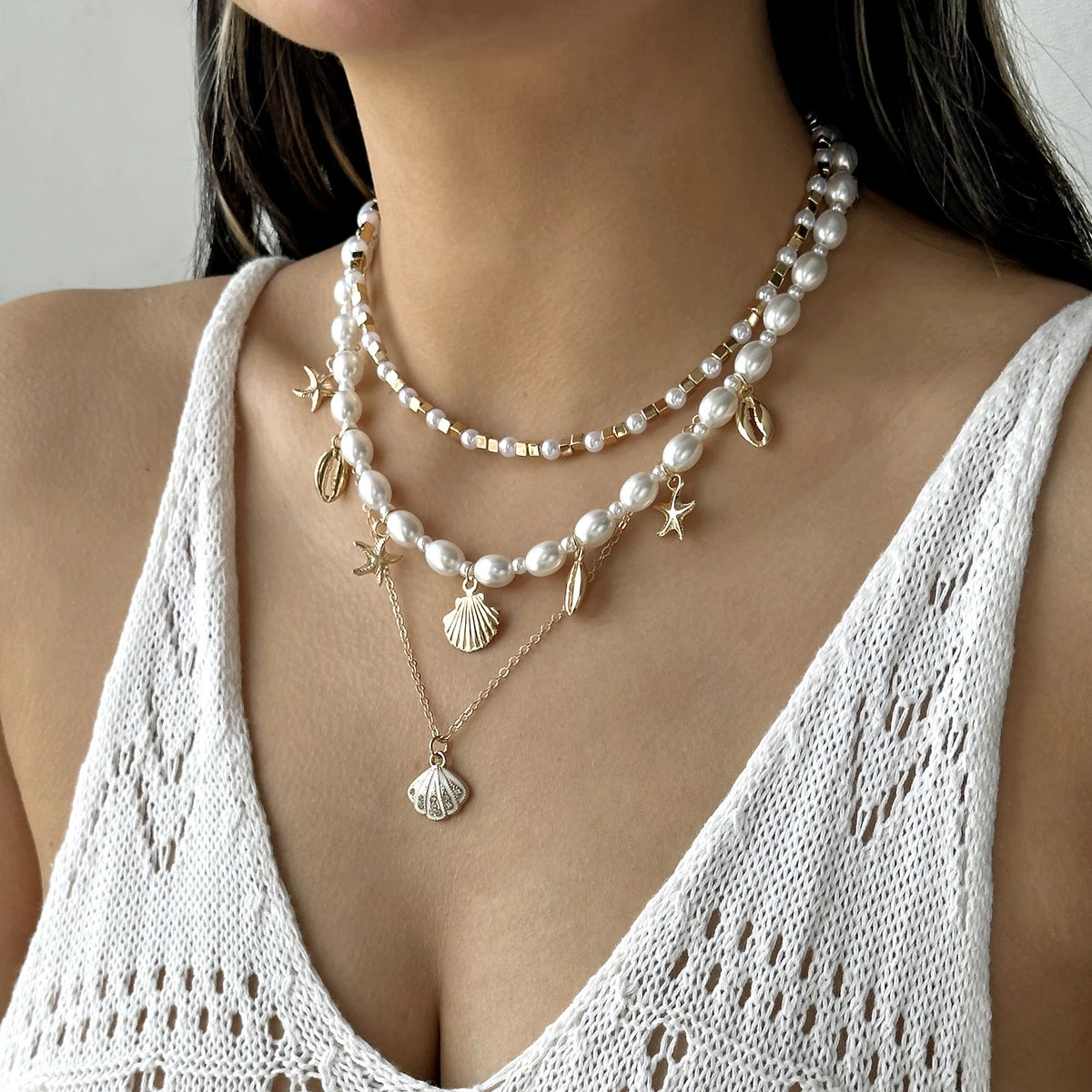 A close-up of a woman wearing a layered necklace featuring pearls, starfish, and shell charms against a white crochet top.