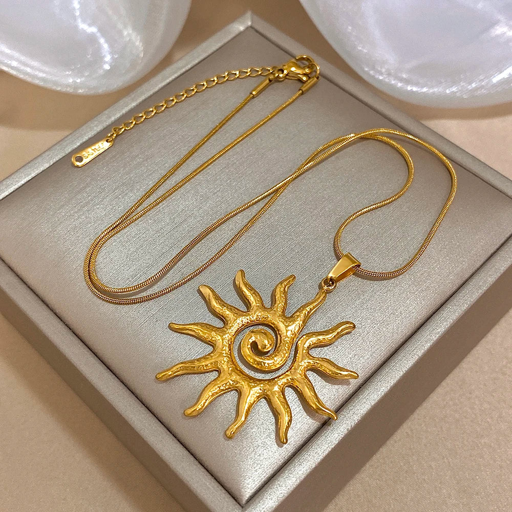 Collier fleur de soleil en spirale plaqué or, présenté sur une boîte carrée avec des éléments décoratifs blancs.