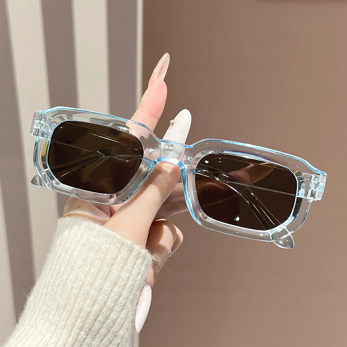 A hand holds a pair of clear acetate retro square sunglasses with dark lenses against a neutral background.