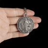 A silver round medallion featuring a depiction of St. Benedict and surrounding text, held in a hand.