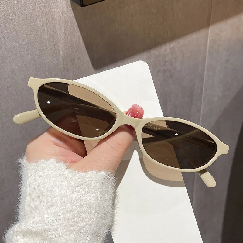 A hand holds a pair of retro oval frame sunglasses with brown lenses against a light background.