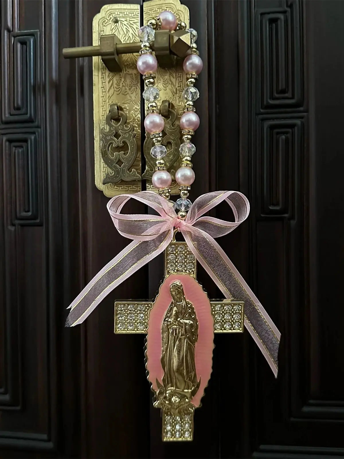 A gold crucifix charm featuring the figure of the Virgin Mary, suspended by a pink ribbon and pearl beads.