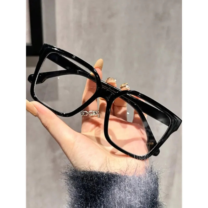 Una mano sostiene unas gafas cuadradas negras anti-luz azul contra un fondo gris claro.