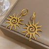 Boucles d'oreilles pendantes en forme de tournesol spiralées plaquées or, présentées sur une boîte de couleur neutre.