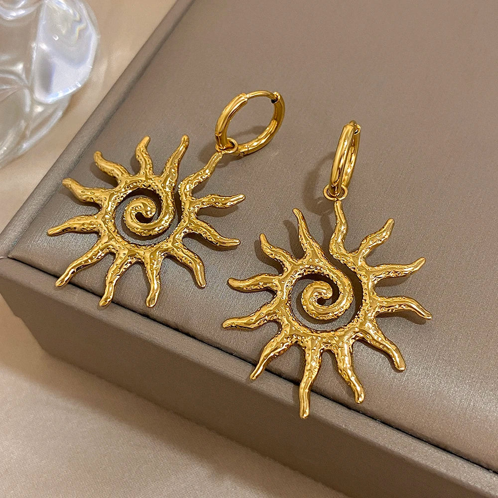 Boucles d'oreilles pendantes en forme de tournesol spiralées plaquées or, présentées sur une boîte de couleur neutre.