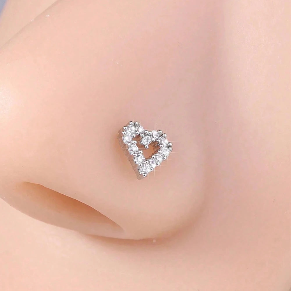 A close-up of a small silver heart horseshoe nose stud earring on a person's nostril.