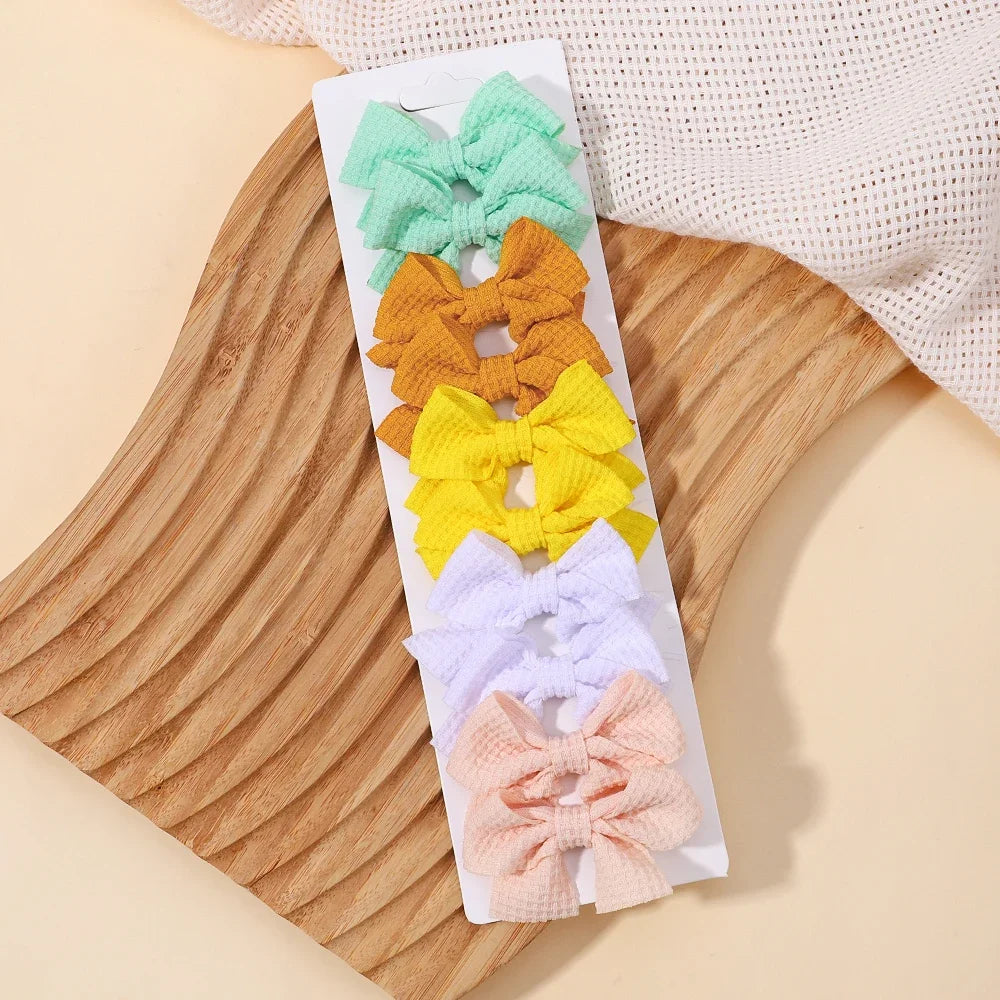 A display of five handmade ribbon bow hair clips in pastel colors including mint green, orange, yellow, white, and pink on a 