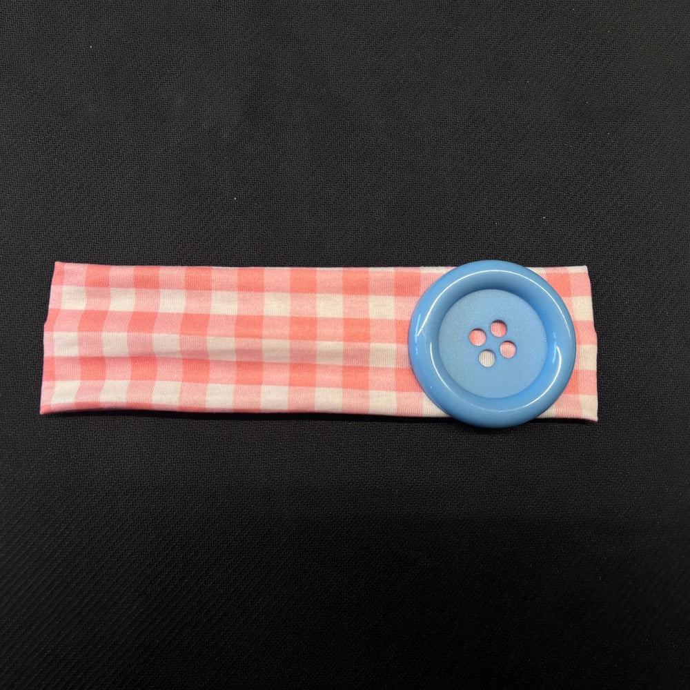 A light pink and white checkered fabric headband with a round blue button attached.
