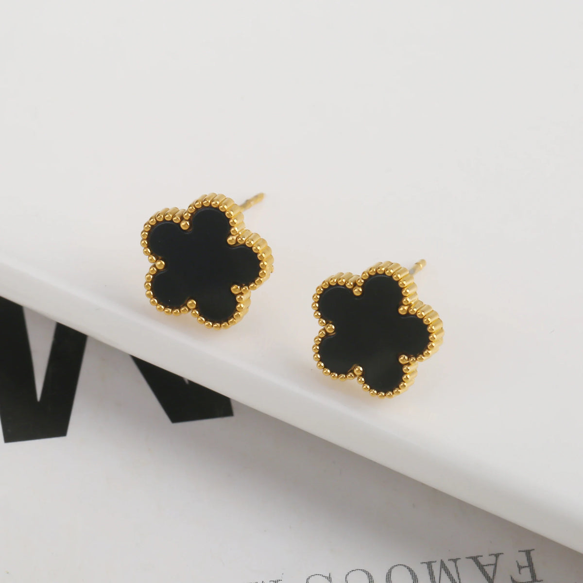 Un par de pendientes florales de planta de acero inoxidable chapado en oro con un diseño negro de cuatro hojas.