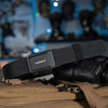 A black tactical nylon belt with a ratchet buckle is shown resting on a tan canvas bag.