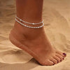 A close-up of a person's ankle showing layered gold-plated twisted bracelets against a sandy background.