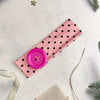 Pink fabric hairband with black polka dots and a pink circular accent on a white background.