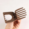 A hand holds a flat circular hair clip set with multiple parallel prongs, colored brown.