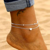 Gros plan sur la cheville d'une femme avec un anklet argenté à perles délicat avec des charms en forme de cœur.
