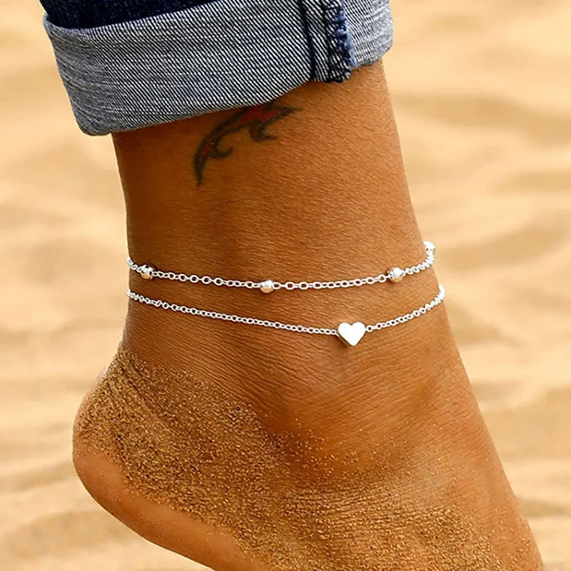 Gros plan sur la cheville d'une femme avec un anklet argenté à perles délicat avec des charms en forme de cœur.