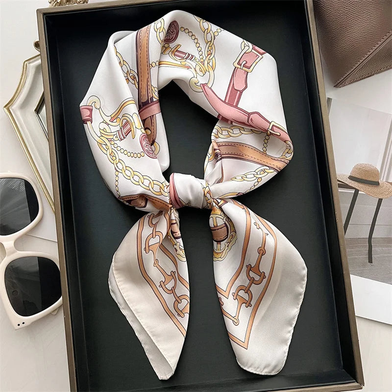 A beige silk scarf tied in a bow, featuring gold and pink embroidery, displayed in a dark tray.