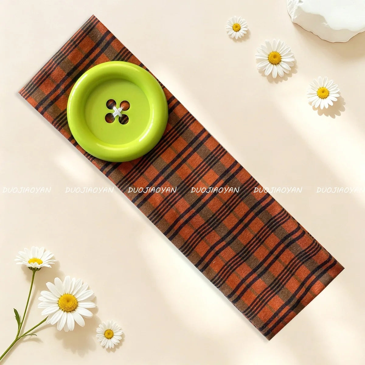 A green button headband rests on a brown plaid fabric with white daisy accents.