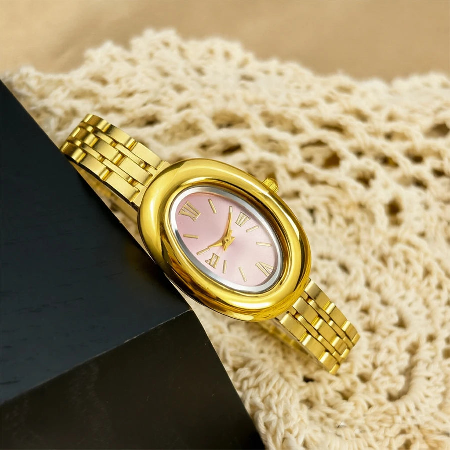 Reloj estilo bañera vintage con esfera rosa y correa dorada sobre tela de ganchillo beige claro.