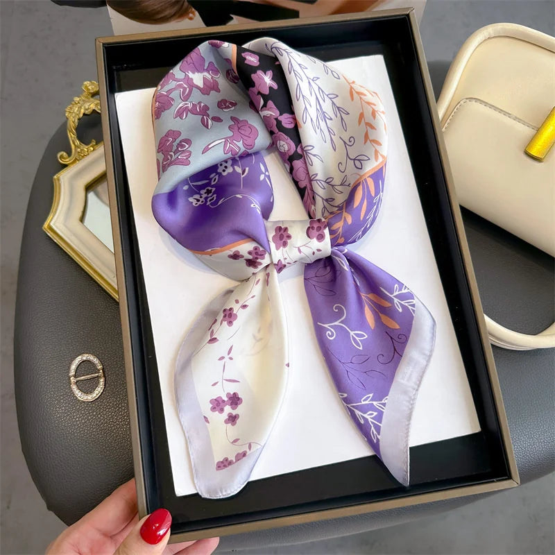 A square silk scarf featuring purple and lilac floral patterns is displayed in a dark box.
