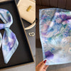 A light blue floral print silk scarf featuring purple and green flowers displayed in a black frame.