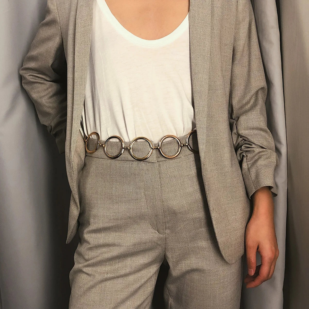 Una persona vistiendo un blazer y pantalones gris claro con un cinturón de cadena metálica dorada.