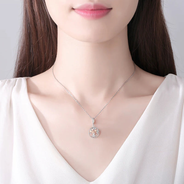 A close-up of a woman wearing a silver necklace with a family tree charm against a white top.