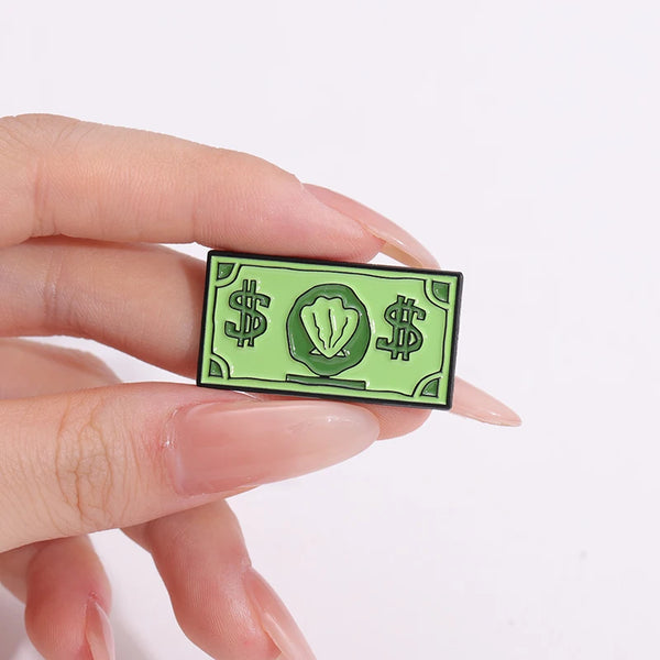 A hand holds a green enamel pin featuring the custom USD symbol and dollar signs.