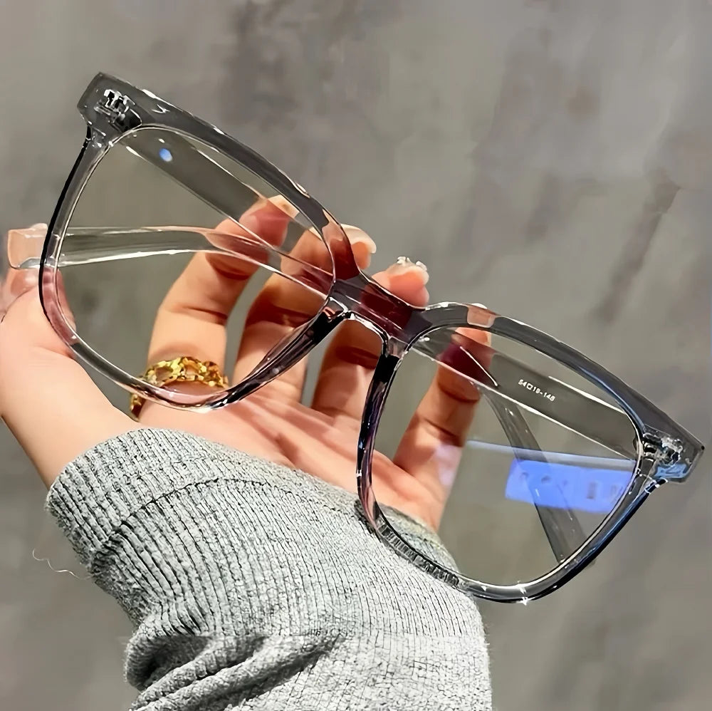 A hand holds a pair of clear, square-shaped eyeglasses with thin purple frames against a gray background.