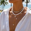 A close-up of a woman wearing a layered white beaded necklace featuring starfish and pearl charms against a bright outdoor ba