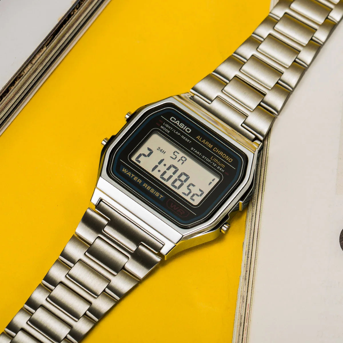 A silver Casio digital wristwatch with a metal bracelet displayed against a bright yellow background.