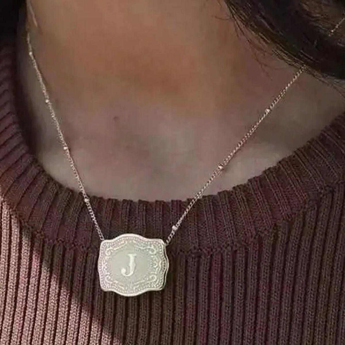 A silver pendant necklace with a carved initial J is displayed against a dark sweater.