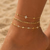 Gold-toned crystal cross anklets set worn on a person's ankle against a sandy background.