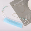 A delicate gold and light blue pearl beaded eyewear chain draped next to a light blue face mask on a gray paper.