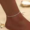 A close-up of a sparkling zircon heart cross anklet worn on a woman's ankle against a sandy background.