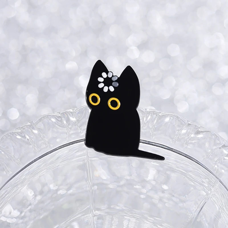 A black cat enamel pin with yellow eyes and white markings resting on a clear glass surface.