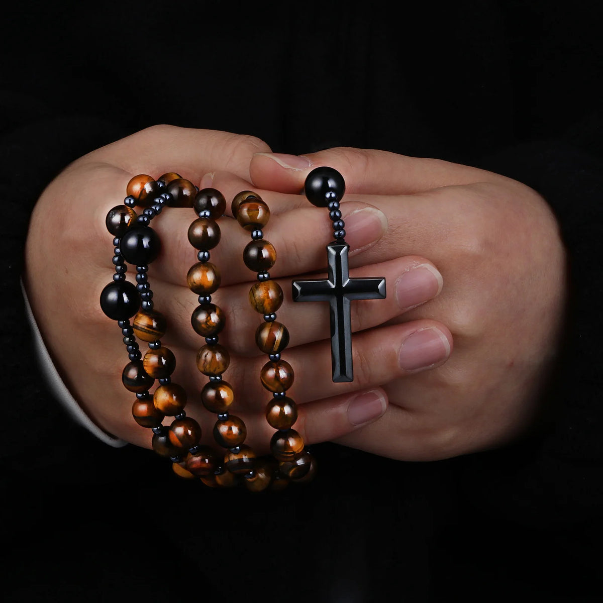 A hand holds a rosary necklace made of tiger eye and black agate beads with a hematite cross pendant.