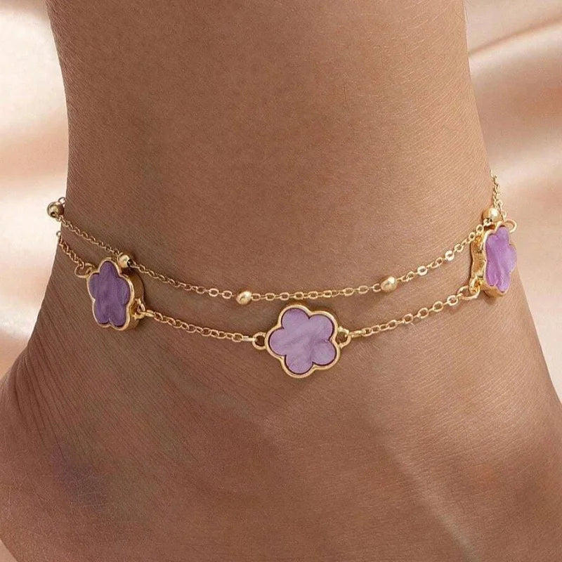 Gold jewelry featuring five-leaf clover anklets with purple stones worn on a person's ankle.