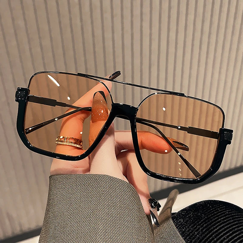 A hand holds a pair of black double bridge oversized square sunglasses with brown tinted lenses against a light background.