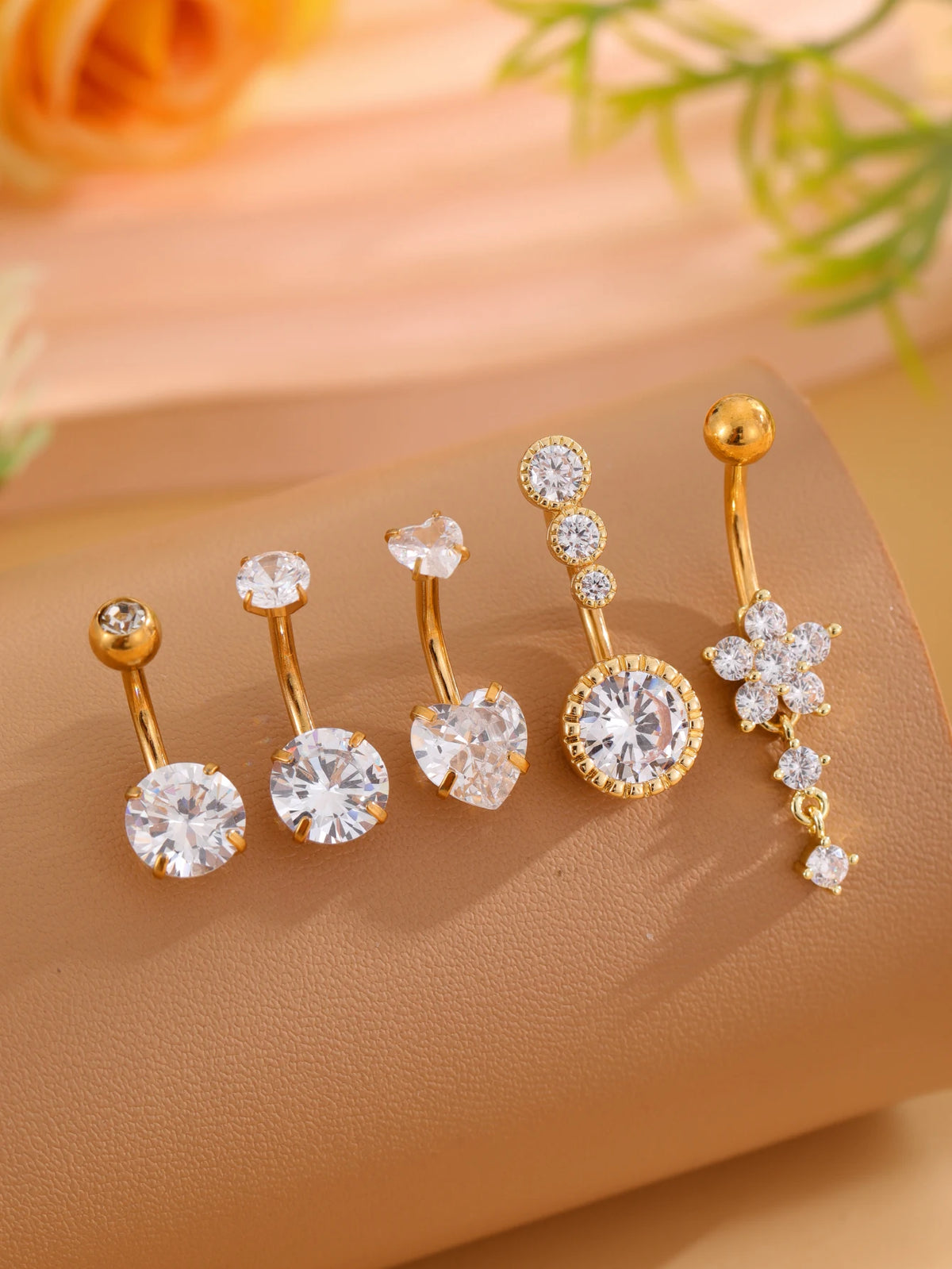 A set of five gold stainless steel dangled belly button rings with various diamond and gemstone designs displayed on a beige 