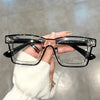 A hand holds a pair of clear square-framed glasses with black arms against a blurred background.